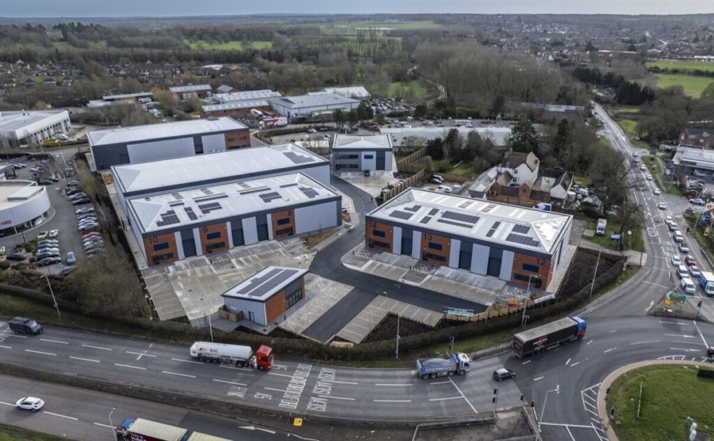 Aerial view of a modern warehouse complex with four long, gray-and-brick buildings numbered 8–14, surrounded by parking lots and access roads in a suburban industrial area, with trucks and cars on the nearby roundabout road."