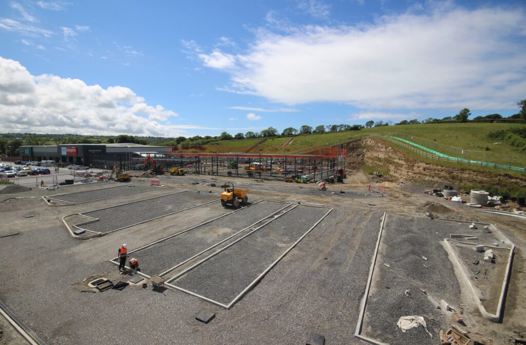 Construction site with workers laying pipes, machinery, and orange safety barriers; a shopping center visible in the background under a blue sky.