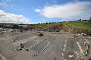 Construction site with workers laying pipes, machinery, and orange safety barriers; a shopping center visible in the background under a blue sky.