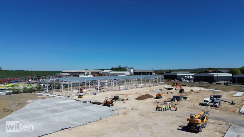 Construction site with a large steel frame structure under construction, workers in high-visibility vests, and parked equipment against a clear blue sky.