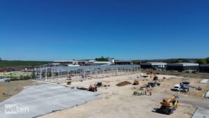 Construction site with a large steel frame structure under construction, workers in high-visibility vests, and parked equipment against a clear blue sky.